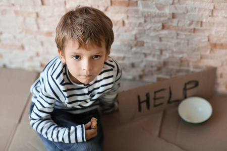 无家可归的可怜的男孩与空的碗和纸箱板用词帮助坐在砖墙附近