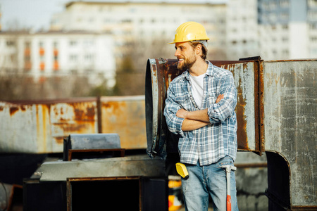 一个强壮的男人, 胡子披着披肩, 头上戴着头盔的工地工人, 用工具, 准备开始工作, 在一个生锈的建筑工地上的某个地方, 站着