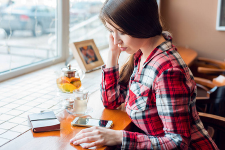 女学生穿着衬衣, 下午在咖啡馆旁边的窗边, 看着电话, 读了一封短信, 茶壶里有一杯茶, 一本笔记本放在一张木桌上。