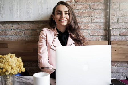 一个美丽的年轻女子的肖像, 穿着粉红色的皮夹克坐在桌子上, 笔记本电脑在咖啡馆里