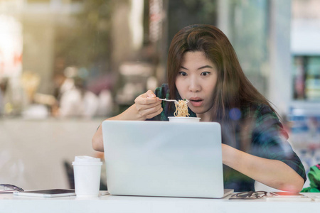 亚洲女商人在休闲西装上的肖像在繁忙时间的办公桌旁, 坐在办公室的玻璃旁边, 工作勤奋的概念, 吃面条与震惊的行动