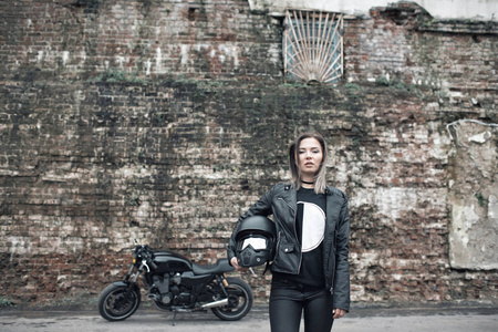 一名骑摩托车的女司机, 手里拿着头盔, 靠近自行车, 车库背景的砖墙。穿着皮夹克和紧身裤的女孩手里拿着摩托车头盔