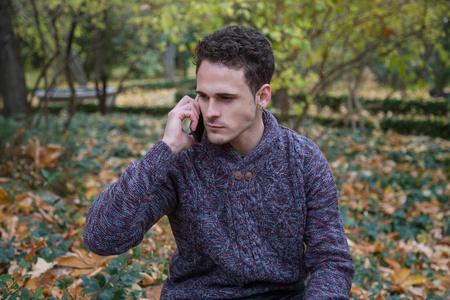 年轻英俊的男子坐在草地上与树叶和通信与智能手机