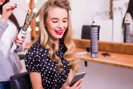 女理发师直发器的长长的头发，微笑的年轻女子，在更衣室里使用智能手机的应用