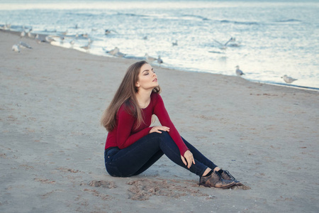 生活方式肖像的白种人年轻美丽的女人闭着眼睛，长长的头发，黑色的牛仔裤和红色的衬衫，坐在沙滩上海滩之间海鸥鸟在秋天落夕阳