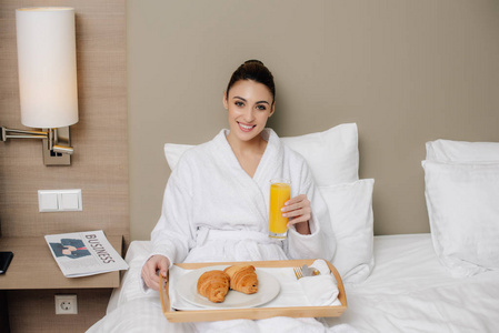 微笑的妇女在浴袍放松在旅馆房间与早餐在床上和看照相机