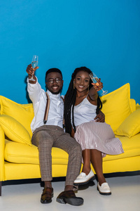 快乐时尚的非洲裔美国人情侣坐在沙发上喝香槟, 微笑着拍照