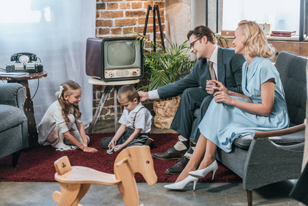 快乐的父母坐在沙发上, 看着小孩子玩多米诺瓷砖在家里, 二十世纪五十年代风格