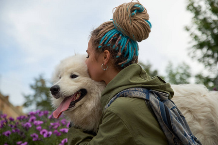 现代时尚的年轻女孩与辫子在她的头拥抱和亲吻她心爱的狗白雪公主萨摩耶。春天公园的背景肖像, 绿色的花朵