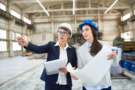 两名女工程师画像 hardhats 站在现代化厂房的宽敞厂房里, 指着建筑规划