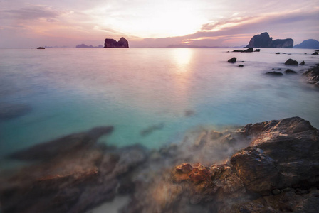 在泰国南部海的倪少杰岛上的岩石海滩上的太阳升起。山背景对五颜六色的红色天空和翡翠蓝绿色的水。慢速快门速度拍摄海景