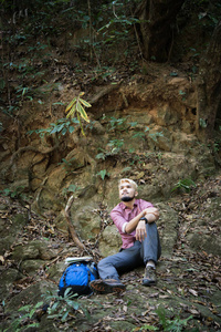 年轻疲惫的徒步旅行者, 背包坐在森林的岩石上, 而在活跃的步行后休息。旅游理念