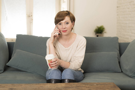 年轻的迷人和快乐的红头发女人坐在家里沙发沙发上喝咖啡谈手机的交流理念放松
