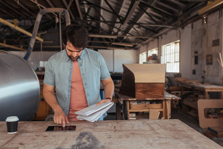 熟练的年轻木工站在他的木工车间工作台使用数字平板电脑和手持设计笔记本