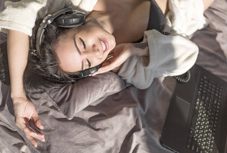 美丽的年轻女子躺在床上, 听音乐从笔记本电脑通过耳机