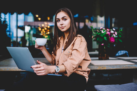 年轻的女商人的肖像在网上赚取金钱使用膝上型电脑, 熟练的女自由职业者做在咖啡馆内部更新软件在咖啡打破看相机的远程工作