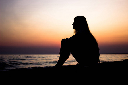 一个年轻女孩的剪影, 坐在沙滩上俯瞰海滩与太阳作为背景。日落
