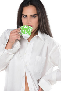 美丽的黑发妇女在白色男性衬衫和内衣拿着一杯咖啡或茶。在白色背景上被隔离