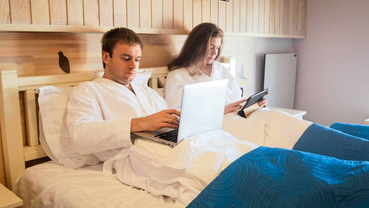 年轻夫妇躺在床上, 并在笔记本电脑和 tablet 计算机上工作