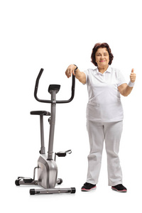一个老妇人的全长画像靠在一辆健身自行车上, 在白色背景上竖起拇指标记。