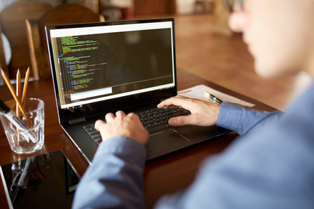 后视图在开发者程序员的肩膀拍摄与笔记本电脑。程序代码和脚本数据在屏幕上。年轻的自由职业者在眼镜工作在咖啡馆的项目