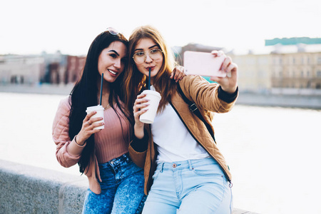在城市背景下旅行时, 最好的朋友会用手里的咖啡制作照片。积极的时髦女孩休息在一起在城市设置和采取自拍图片的智能手机