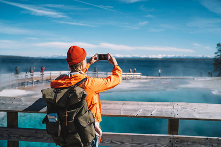 女性旅行者的背部视图使用智能手机相机拍照的温泉, 时髦的女孩与背包拍照片进入黄石公园旅游地标在美国之行