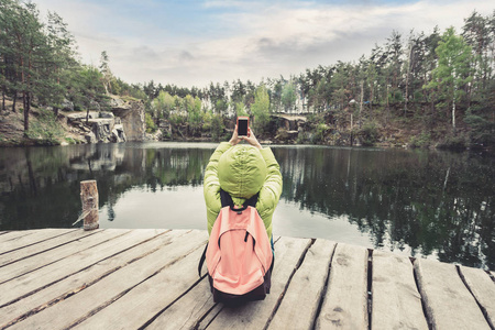 女性旅行者坐在附近一个美丽的湖，在一片松树林和拍照手机上的木码头上