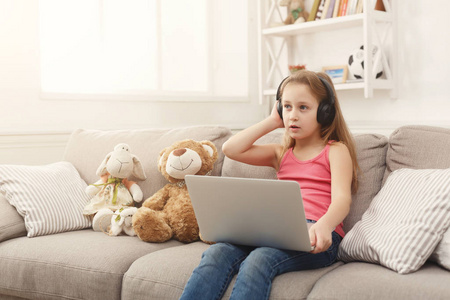 可爱的小女孩在笔记本电脑上做作业, 坐在沙发上戴着耳机