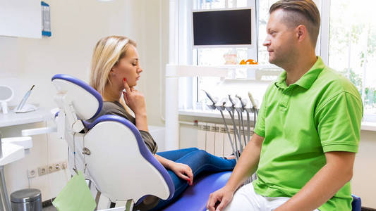 年轻的金发女郎在牙科椅上, 并显示疼痛的牙齿给她的医生