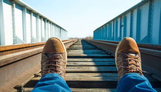 棕色皮革运动鞋和蓝色男性牛仔裤。脚超过铁轨。铁路上的男子脚运动鞋
