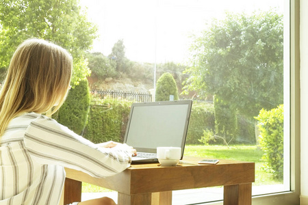漂亮的时髦女人穿着宽松条纹衬衣坐在家里, 用个人计算机。女作家在家里写剧本, 靠大窗户, 喝咖啡。复制空间背景特写
