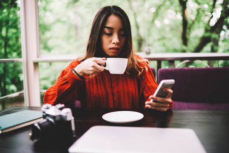 沉思的亚洲时髦的女孩享受热咖啡休息在咖啡馆, 在网上商店购买付款在手机上应用, 年轻体贴的女人在手机上更新软件在休息期间