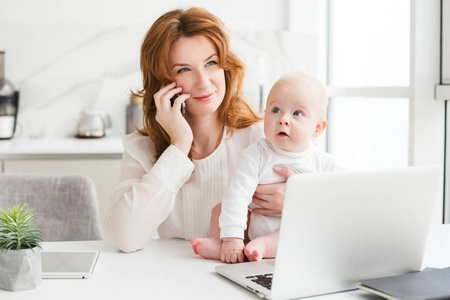 微笑的商业妇女的肖像坐在桌子上与笔记本电脑, 并在她的手机交谈, 同时抱着她可爱的小婴儿在手隔离