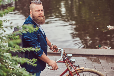有胡子的男性与一个时髦的发型穿着休闲服装与背包, 站在河边的复古自行车在城市公园