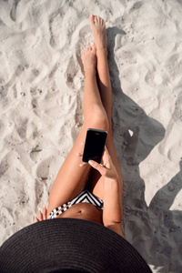 女孩使用手机应用程序短信短信或浏览在线社交媒体。夏天海滩假期。穿着遮阳帽的妇女在海滩上放松