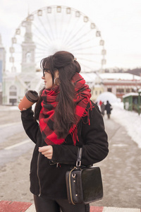 可爱的年轻白种黑发女学生在欧洲城市街道散步。她穿着拉丁大衣和格子红围巾。寒冷的冬天天气。妇女喝咖啡从一个到去的杯子