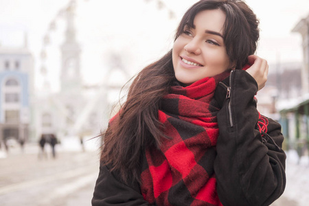 可爱的年轻白种黑发女学生在欧洲城市街道散步。她穿着拉丁大衣和格子红围巾。寒冷的冬天天气