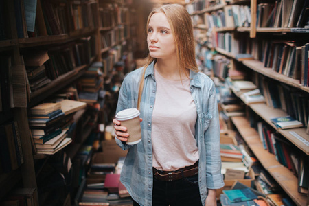 有魅力和善良的女孩站在大而长的 bookshelfs 与旧书。她手里拿着一杯咖啡, 看着架子。那个女人正在找一本书。