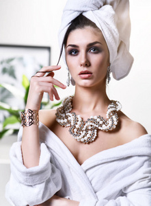 年轻美丽的女人用白色的毛巾头上看着镜子穿着浴衣和金珍珠首饰