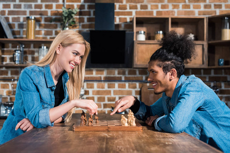 微笑的多民族年轻夫妇在厨房一起下棋的侧面视图