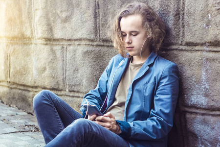 一个十几岁的男孩听音乐, 在城市环境中使用电话。一个英俊的小伙子坐在人行道上写短信