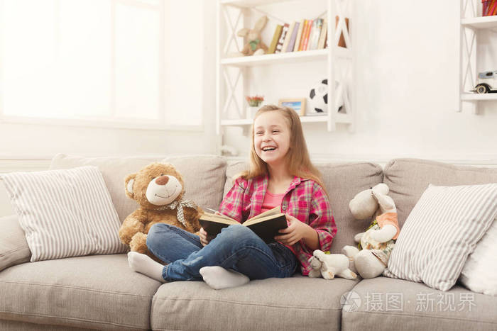 快乐的小女孩拥抱她的泰迪熊和读书在沙发在家
