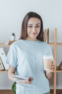 美丽的微笑的女孩拿着抄写本和咖啡杯