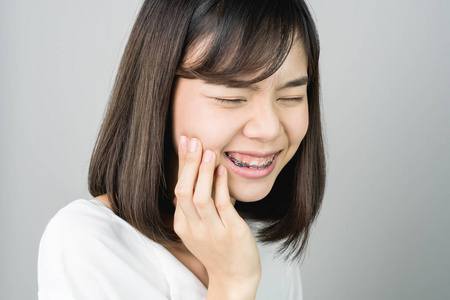 亚洲女孩穿着白色休闲服装炫耀牙痛, 也许是因为没有保持良好的口腔健康。在灰色背景下给出柔和的光