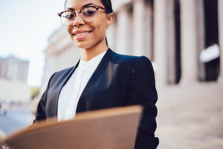 美国黑人女商人在镜头中微笑的裁剪图像。成功的女性企业雇员站在城市设置附近的办公大楼与文件文件夹在手