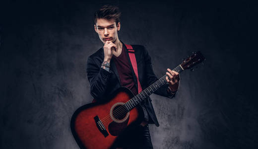 英俊的年轻体贴的音乐家与时髦的头发穿着优雅的衣服摆在他的手中的吉他