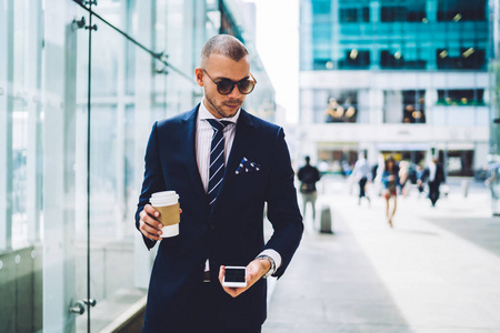 英俊的男性企业家检查在智能手机上的邮件, 而步行在市中心与咖啡去, 认真的商人在太阳镜下城市金融区移动行走的信息传递