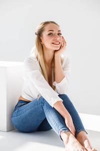 金发女郎的肖像穿着蓝色牛仔裤和白色衬衫坐在白色工作室背景