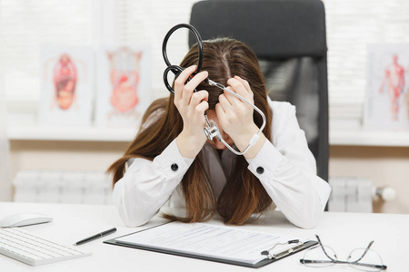 年轻疲惫的疲惫的妇女坐在办公桌前, 在电脑上工作, 在医院的光办公室的医疗文件。女医生在医疗礼服睡在咨询室。保健医学概念